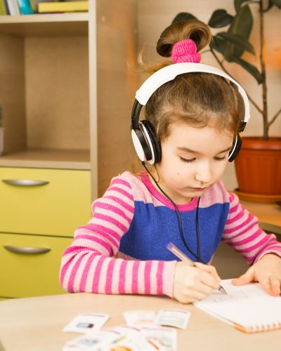 A pre-school girl with headphones is engaged at home at the table studying an educational program, writing in a notebook. Distance learning, education, language learning. Copy space